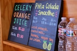 Two black chalkboards list drink options: one offers celery orange juice, and the other features a piña colada smoothie. Prices and sizes are displayed in vibrant colors. Two plastic water bottles sit beside the boards, partially visible.