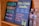 Two black chalkboards list drink options: one offers celery orange juice, and the other features a piña colada smoothie. Prices and sizes are displayed in vibrant colors. Two plastic water bottles sit beside the boards, partially visible.