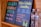 Two black chalkboards list drink options: one offers celery orange juice, and the other features a piña colada smoothie. Prices and sizes are displayed in vibrant colors. Two plastic water bottles sit beside the boards, partially visible.