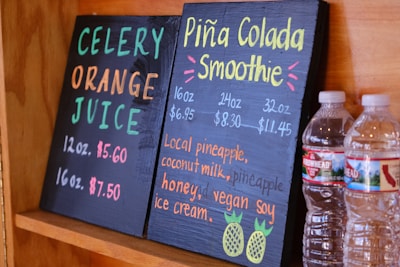 Two black chalkboards list drink options: one offers celery orange juice, and the other features a piña colada smoothie. Prices and sizes are displayed in vibrant colors. Two plastic water bottles sit beside the boards, partially visible.