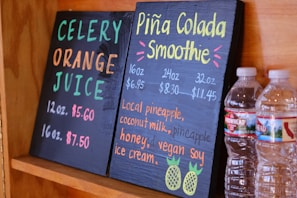 Two black chalkboards list drink options: one offers celery orange juice, and the other features a piña colada smoothie. Prices and sizes are displayed in vibrant colors. Two plastic water bottles sit beside the boards, partially visible.