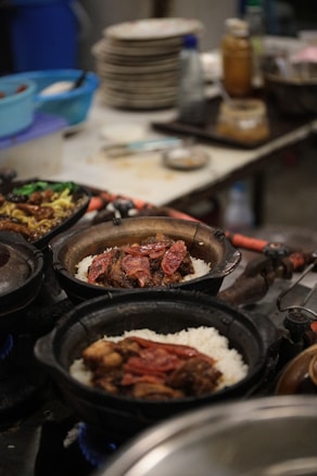 A kitchen scene features clay pots on a stove with rice and sliced meat, possibly sausage. In the background, there are stacks of plates, various utensils, and containers. The setting appears busy and functional, suggesting a restaurant kitchen environment.