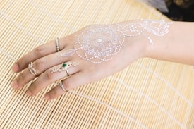 An elegantly adorned hand rests on a bamboo mat. The hand features intricate white henna designs with floral and paisley patterns on the skin. Several ornate rings embellish the fingers, including one with a prominent green gemstone.