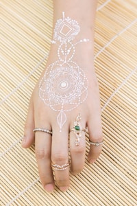 A hand adorned with intricate white henna designs that extend from the wrist onto the back of the hand. Several decorative rings with gemstones, including one with a green stone, embellish the fingers. The hand rests on a bamboo mat, providing a natural, textured background.