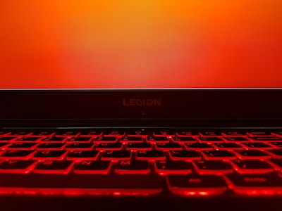 A laptop keyboard illuminated with a bright red backlight is in the foreground, while the screen displays a plain orange background. The brand name 'LEGION' is visible above the keyboard.