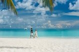 A sunlit beach with turquoise waves gently meeting white sand and a couple walking hand in hand.