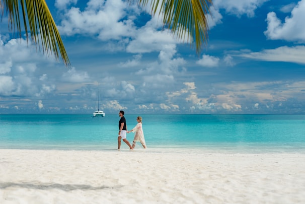 A sandy beach with turquoise waves gently rolling in and a couple walking hand in hand.