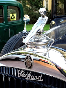 A vintage Packard car hood ornament, depicting a sleek, stylized figure, is prominently displayed. The emblem is shiny with intricate details, set against the polished chrome grille. To the side, a green car with classic design elements can be seen, parked on a grassy area with trees in the background.