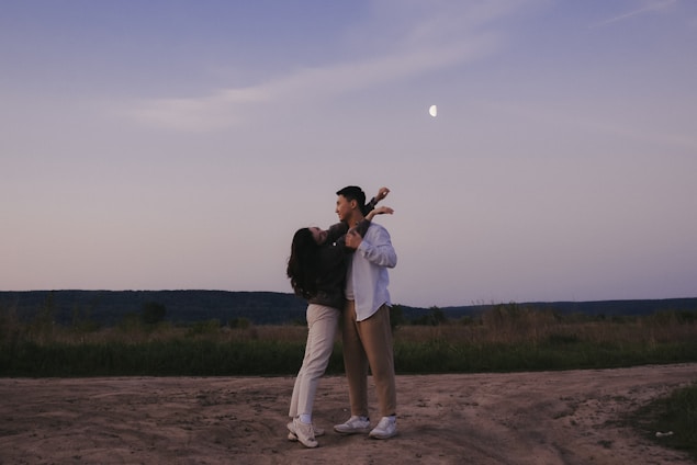 A couple stands on an open dirt path, embraced in a dance under a dusky sky. The moon is visible in the background, partially illuminating the scenic landscape around them.
