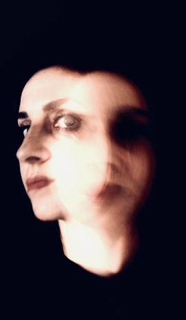 A blurred portrait of a person's face with overlapping images, creating a sense of motion or duality. The lighting is dramatic, highlighting the facial features against a dark background.