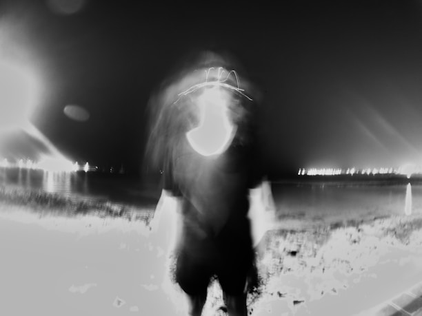 A blurred, monochromatic image featuring a ghostly figure standing in the foreground against a nighttime backdrop. The background includes a body of water with distant lights reflecting on its surface, creating an eerie and surreal atmosphere.