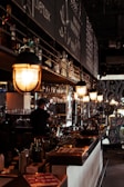A cozy, dimly lit bar interior featuring hanging lights above the counter, various bottles of alcohol on shelves, and a person working behind the bar. Chalkboard menus are visible in the background with writing detailing drinks or specials.