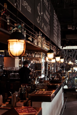A cozy, dimly lit bar interior featuring hanging lights above the counter, various bottles of alcohol on shelves, and a person working behind the bar. Chalkboard menus are visible in the background with writing detailing drinks or specials.