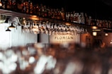A dimly lit bar scene features hanging glasses and shelves stocked with various bottles of liquor. Warm lighting from antique-style lamps casts a cozy glow over the counter. The name 'Florian' is visible on the back wall, suggesting the name of the establishment.