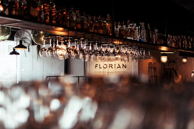 A dimly lit bar scene features hanging glasses and shelves stocked with various bottles of liquor. Warm lighting from antique-style lamps casts a cozy glow over the counter. The name 'Florian' is visible on the back wall, suggesting the name of the establishment.