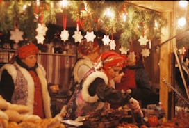 Women dressed in traditional festive attire are serving food at a market stall adorned with holiday decorations. The setting includes pine garlands and white snowflake ornaments hanging above. The scene is warmly lit, creating a cozy atmosphere.