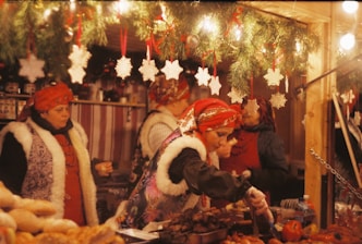 Women dressed in traditional festive attire are serving food at a market stall adorned with holiday decorations. The setting includes pine garlands and white snowflake ornaments hanging above. The scene is warmly lit, creating a cozy atmosphere.