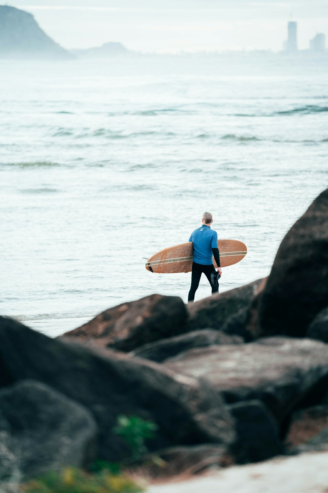 man in blue shirt and black pants holding brown surfboard standing on gray rock near body