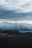 A telecommunications tower rises above a cityscape under a thick, overcast sky. The clouds appear heavy and dark, creating a moody atmosphere. The foreground features rooftops with some industrial structures and antennas.