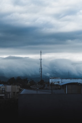A telecommunications tower rises above a cityscape under a thick, overcast sky. The clouds appear heavy and dark, creating a moody atmosphere. The foreground features rooftops with some industrial structures and antennas.