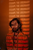 A man stands with a projection of the phrase 'Escape the Ordinary' repeatedly displayed across his face and body. The background appears dimly lit with a warm, reddish hue, creating a striking visual effect.