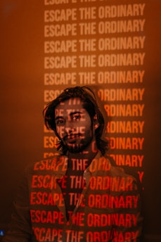 A man stands with a projection of the phrase 'Escape the Ordinary' repeatedly displayed across his face and body. The background appears dimly lit with a warm, reddish hue, creating a striking visual effect.
