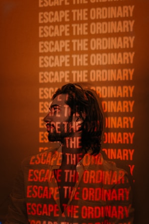 A person is standing in front of a wall covered with the repeated phrase 'Escape the Ordinary' in bold, red letters. The phrase is projected onto the person's shirt and face, creating an overlay effect. The background and ambient lighting are predominantly orange and muted.