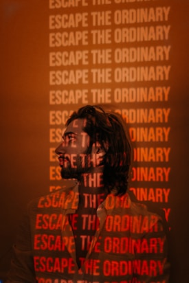 A person is standing in front of a wall covered with the repeated phrase 'Escape the Ordinary' in bold, red letters. The phrase is projected onto the person's shirt and face, creating an overlay effect. The background and ambient lighting are predominantly orange and muted.