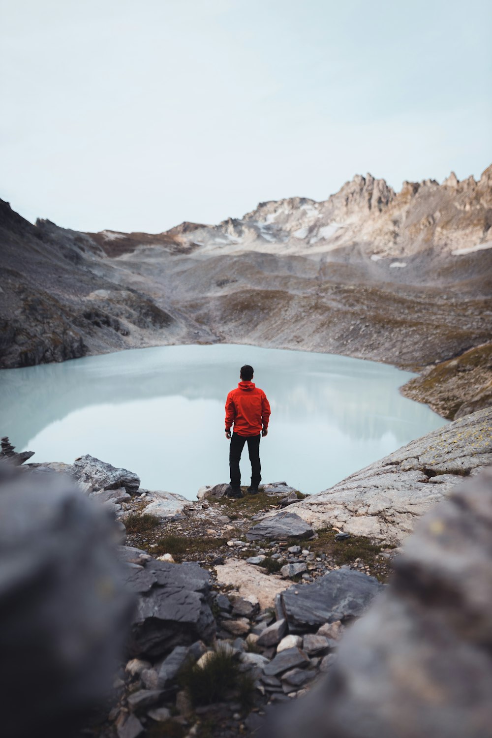 uomo in giacca rossa in piedi su terreno roccioso vicino al lago durante il giorno