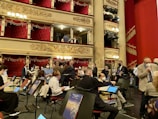 A group of people, some wearing masks, are gathered in an ornate theater setting with tiered balconies and red velvet curtains. Music stands and chairs are positioned on what appears to be a stage area. The theater is lavishly decorated with gold embellishments and hanging chandeliers.