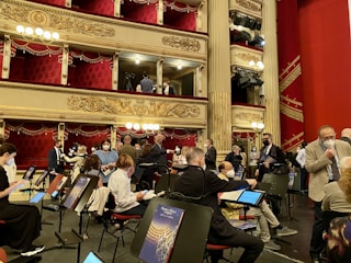 A group of people, some wearing masks, are gathered in an ornate theater setting with tiered balconies and red velvet curtains. Music stands and chairs are positioned on what appears to be a stage area. The theater is lavishly decorated with gold embellishments and hanging chandeliers.