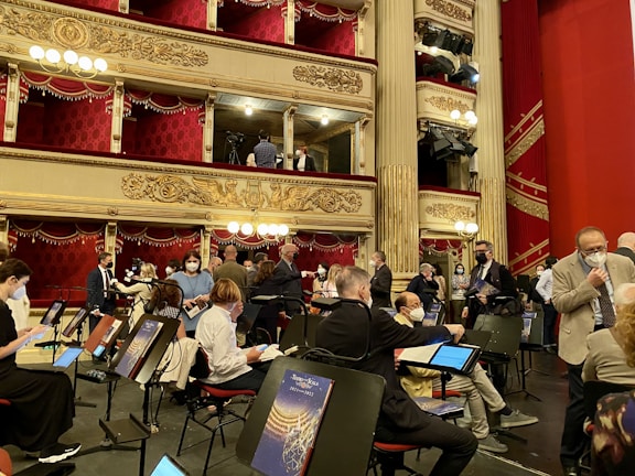 A group of people, some wearing masks, are gathered in an ornate theater setting with tiered balconies and red velvet curtains. Music stands and chairs are positioned on what appears to be a stage area. The theater is lavishly decorated with gold embellishments and hanging chandeliers.