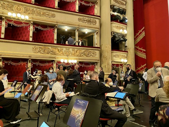 A group of people, some wearing masks, are gathered in an ornate theater setting with tiered balconies and red velvet curtains. Music stands and chairs are positioned on what appears to be a stage area. The theater is lavishly decorated with gold embellishments and hanging chandeliers.