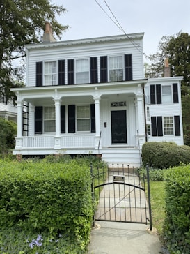 A two-story white house with a classic design, featuring black shutters on the windows, a covered front porch with white railings, columns, and a black front door. The house is surrounded by lush green bushes, and there is a small garden area in front of the porch. A black metal gate at the entrance has a sign that reads 'Private Residence'. Trees and other houses are visible in the background under a slightly overcast sky.