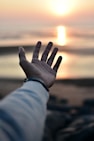 a hand reaching out towards the ocean at sunset