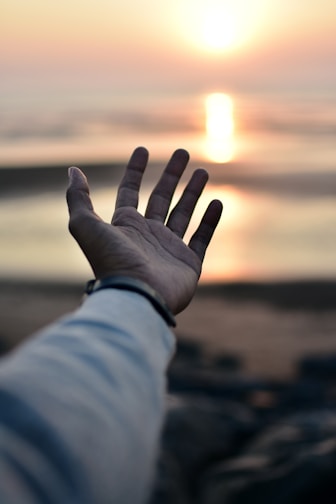 a hand reaching out towards the ocean at sunset