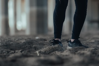 Close-up of a person's legs wearing black leggings and sneakers, standing on a sandy surface. The background appears blurred, suggesting an urban or industrial setting.