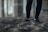 Close-up of a person's legs wearing black leggings and sneakers, standing on a sandy surface. The background appears blurred, suggesting an urban or industrial setting.