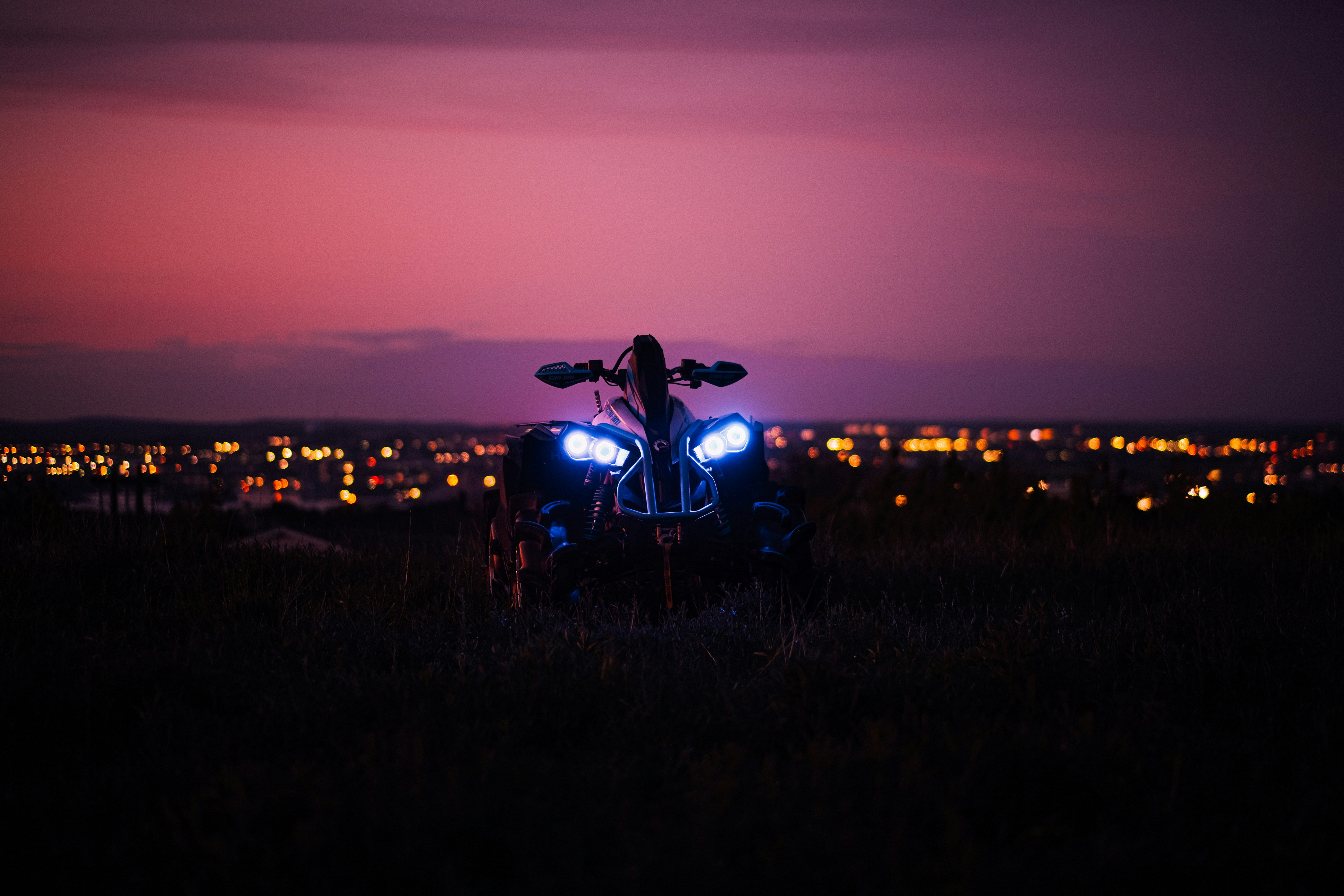 Headlights of an ATV in front of a pink and purple sunset sky and lights of a cityy