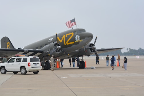 An old military aircraft is displayed on a tarmac with an American flag on top. Several people are gathered around the plane, including some in military uniforms. A white SUV is parked nearby, and there are orange safety cones on the ground.