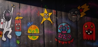 A colorful graffiti mural featuring a variety of cartoon-like characters, including an astronaut, a cylindrical figure with an expressive face, a planet with a ring and greenery, a large yellow star with a face, a small planet or spaceship with a heart, a dark cloud with a surprised face, and a red, ghost-like creature with sharp teeth. The background is a dark, wooden surface, adding contrast to the vibrant figures.