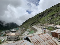 A mountainous landscape with a winding road that traverses through a lush, green valley. The sky is partly cloudy, and mist shrouds the peaks, creating a serene atmosphere. Rustic buildings with corrugated roofs dot the area, and a wooden gazebo stands in the foreground, offering a picturesque view.
