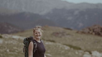 A close-up of a smiling traveler wearing a backpack with a scenic mountain range in the background.