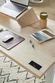 A minimalist workspace with a wooden desk, featuring a laptop on a wooden stand, a closed book or notebook with abstract art on its cover, a lit candle in a two-toned beige and green holder, a wireless mouse on a leather pad, two green dice, and a portion of a patterned rug visible in the corner.