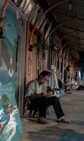 An artist is sitting on a small stool in a covered corridor, intently working on a canvas. The area is lined with paintings and framed artworks hung on the walls. The artist is wearing a casual outfit and a face mask, and the atmosphere appears relaxed and focused.