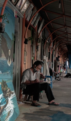 An artist is sitting on a small stool in a covered corridor, intently working on a canvas. The area is lined with paintings and framed artworks hung on the walls. The artist is wearing a casual outfit and a face mask, and the atmosphere appears relaxed and focused.