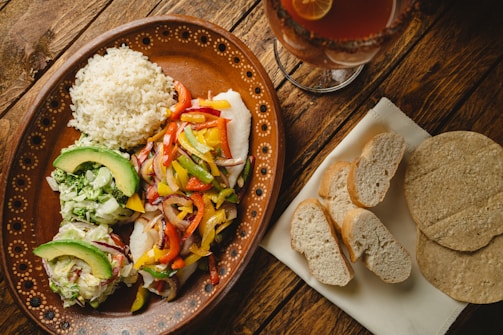 A rustic wooden table displays a vibrant, traditional meal on an ornate brown ceramic plate. The dish includes a serving of white rice, colorful sautéed bell peppers, and onions, alongside avocado slices and mixed greens. A separate napkin holds slices of bread and corn tortillas. A glass of a dark red beverage garnished with a slice of lime is also visible.