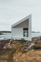 A modern, minimalist structure with an angular, geometric design is situated by a rocky shoreline. The building has a white exterior with a slanted roof and large window. A stack of firewood is placed nearby on the grassy ground, which is dotted with rocks. The background features a body of water and distant landforms under an overcast sky.
