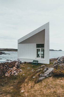 A modern, minimalist structure with an angular, geometric design is situated by a rocky shoreline. The building has a white exterior with a slanted roof and large window. A stack of firewood is placed nearby on the grassy ground, which is dotted with rocks. The background features a body of water and distant landforms under an overcast sky.