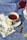 A white teacup filled with tea is placed on a saucer, with a slice of lemon and a cinnamon stick nearby. The setup is on a decorative blue tablecloth with floral patterns. A floral-patterned napkin is folded next to the cup. Red and pink roses in a white vase are partially visible on the right.